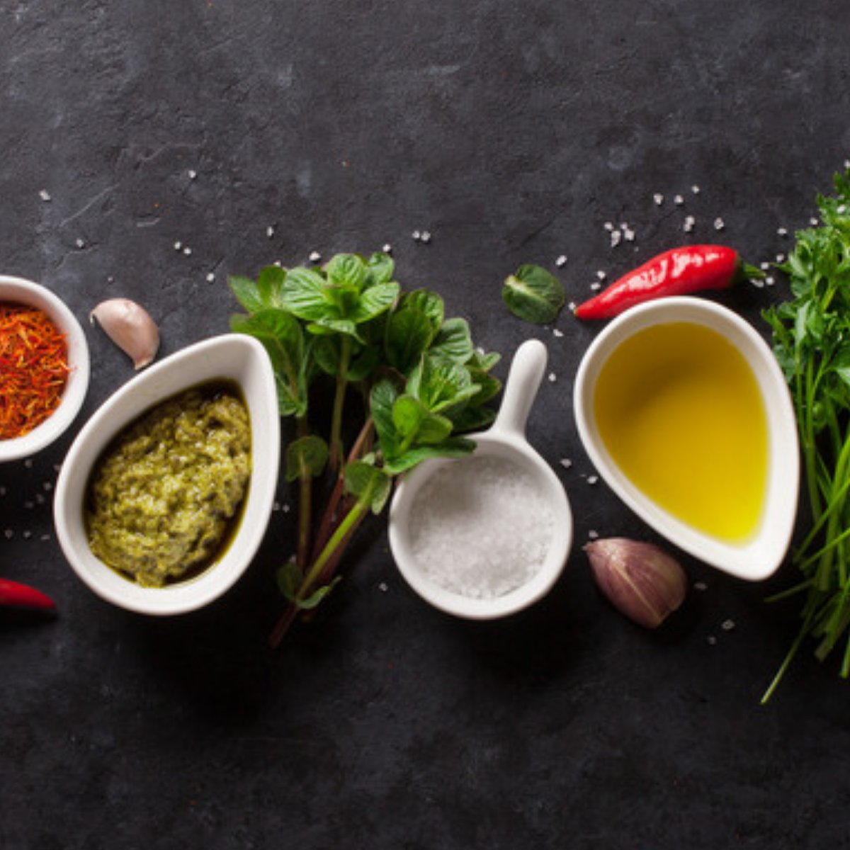 Sauce dishes with olive oil, pesto, salt, chillies and fresh herbs on dark slate