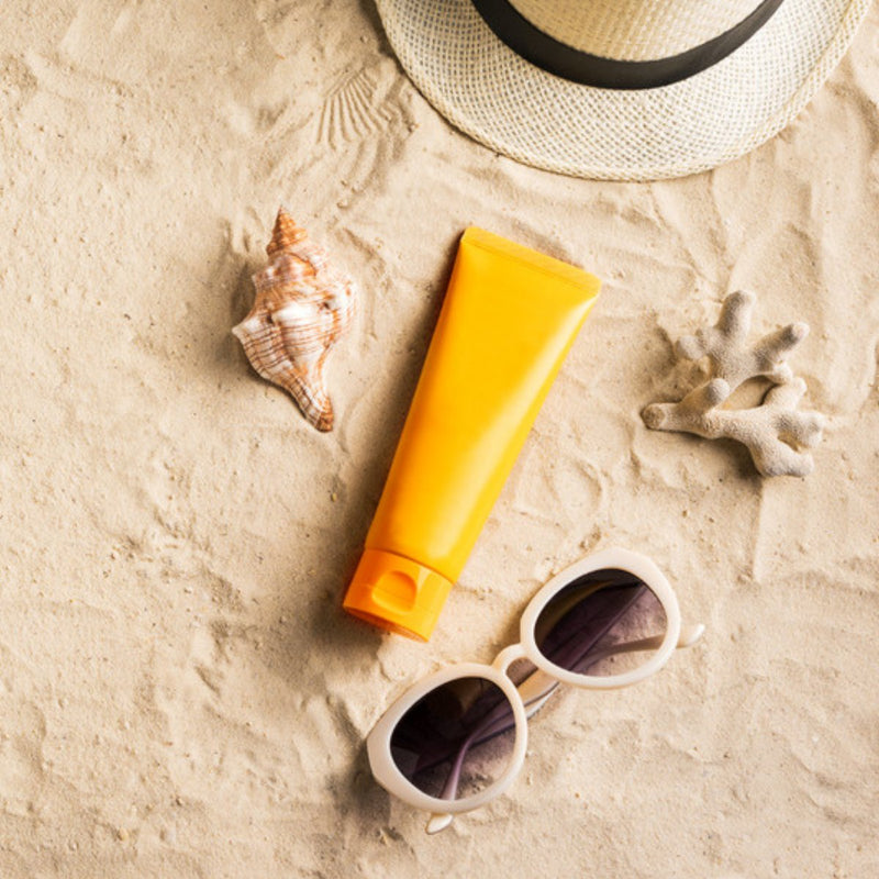 Orange sunscreen tube with sunglasses, shell and straw hat on sand